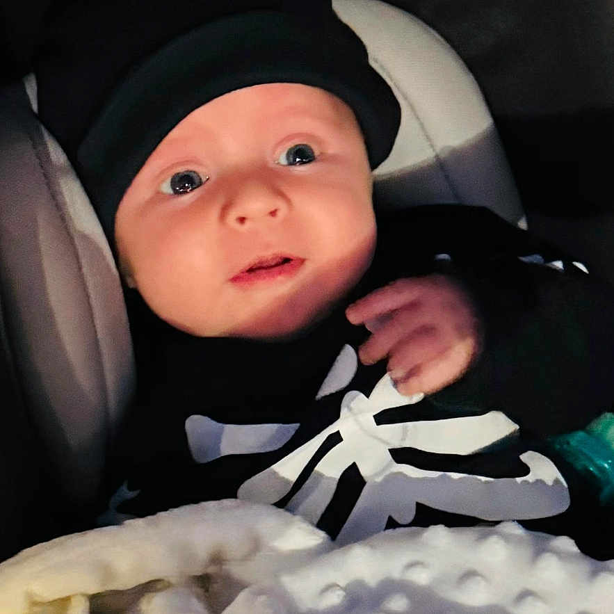 Lukas E joined the competition — help win amazing prizes! baby, black_clothing, blanket, child, clothing, cozy, cute, dark_background, expression, face, hat, indoors, infant, newborn, person, portrait, sitting, soft_texture, warm, wrapped