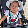 toddler, child, stroller, hat, white_hat, harness, outdoor, sunny, curious_expression, white_tshirt, colorful_design, baby, seatbelt, seat, person, infant, daylight, casual_clothing, summer, portrait