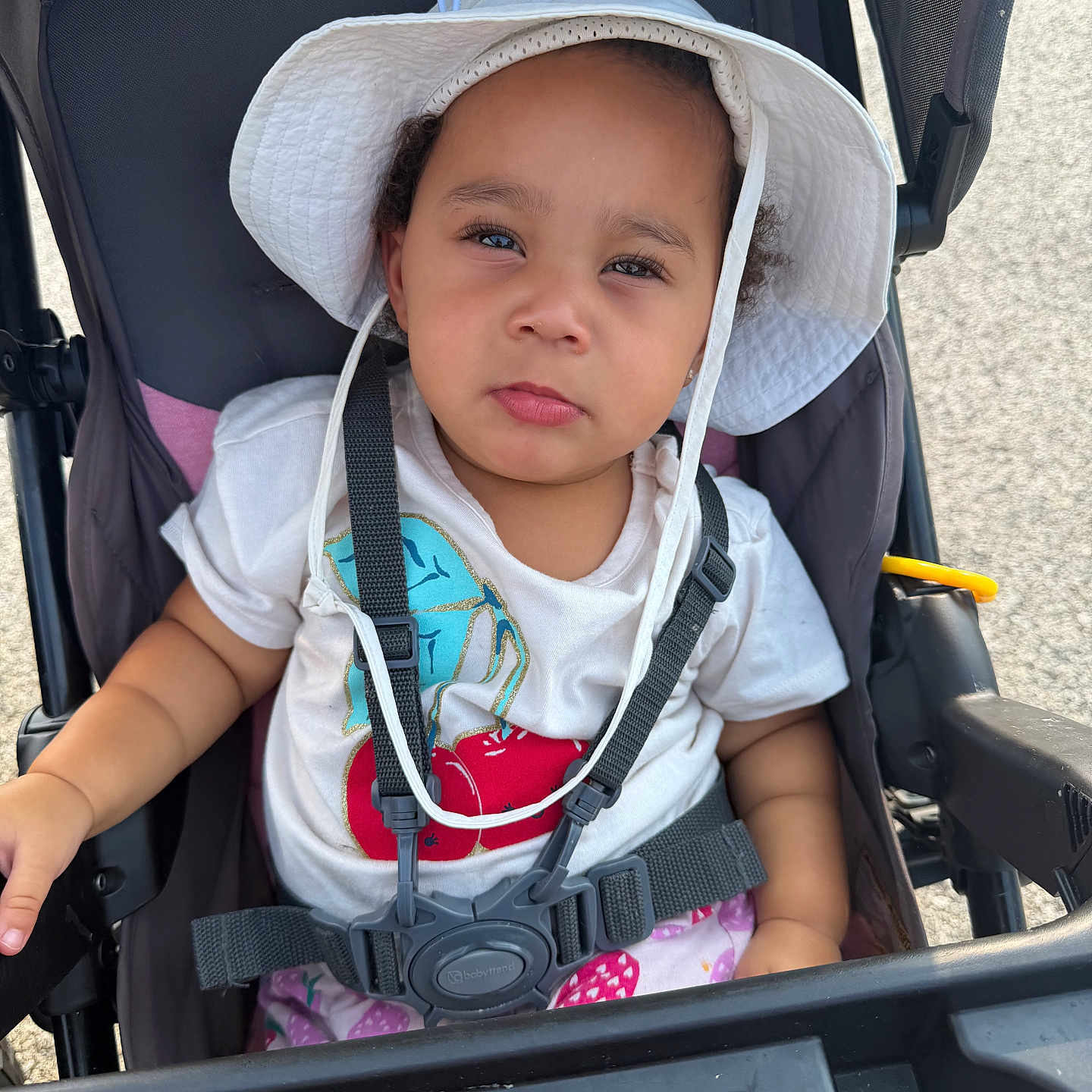 Nehlani is registered to the contest to win money with this photo: toddler, child, stroller, hat, white_hat, harness, outdoor, sunny, curious_expression, white_tshirt, colorful_design, baby, seatbelt, seat, person, infant, daylight, casual_clothing, summer, portrait