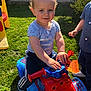 Paddy is registered to the contest to win money with this photo: bare_arms, blue, blue_shirt, casual_clothing, child, curly_hair, dinosaur_print, fun, garden, grass, outdoor, person, plastic_toy, play, red, ride_on_toy, smiling, sunlight, toddler, toy_vehicle