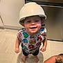 adult_hand, barefoot, captain_america, child, clothing, construction_helmet, floor_tiles, happy, helmet, indoor, kitchen, looking_up, person, playful, shorts, smile, spiderman, superhero_shirt, tattoo, toddler