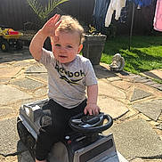 Paddy joined the competition — help win amazing prizes! angel_statue, barefoot, casual_clothing, child, clothes, curly_hair, fence, garden, grass, hand_wave, laundry, outdoor, pavement, potted_plant, smiling, steering_wheel, sunny, toddler, toy_car, toy_truck