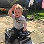 angel_statue, barefoot, casual_clothing, child, clothes, curly_hair, fence, garden, grass, hand_wave, laundry, outdoor, pavement, potted_plant, smiling, steering_wheel, sunny, toddler, toy_car, toy_truck