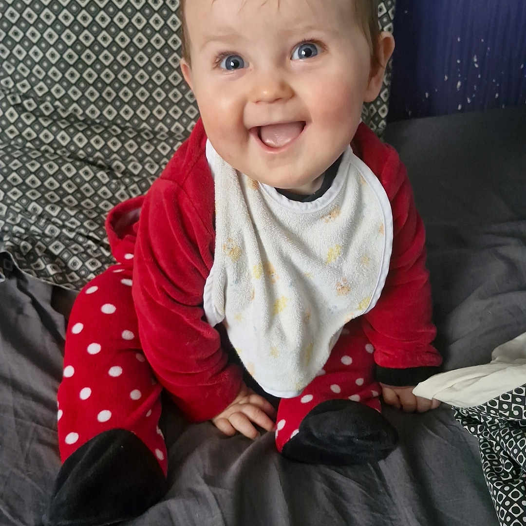 Kléo a rejoint le concours — aidez-le/la à gagner de superbes lots ! baby, bed, bib, blanket, blue_eyes, child, cozy, cute, face, happy, human, indoors, infant, pajamas, patterned_pillow, pillow, polka_dot, red_clothing, sitting, smiling