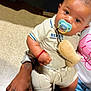 baby, pacifier, plush_toy, hand, watch, ring, bag, clothing, floor, person, lap, tattoo, skin, footwear, jeans, sitting, cute, indoor, accessory, bracelet