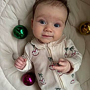 Oli is registered to the contest to win money with this photo: baby, infant, face, person, clothing, onesie, bed, quilt, ornament, christmas_bauble, green, pink, gold, expression, cute, looking_at_camera, indoor, child, small_hands, soft_lighting