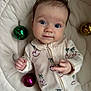 baby, infant, face, person, clothing, onesie, bed, quilt, ornament, christmas_bauble, green, pink, gold, expression, cute, looking_at_camera, indoor, child, small_hands, soft_lighting