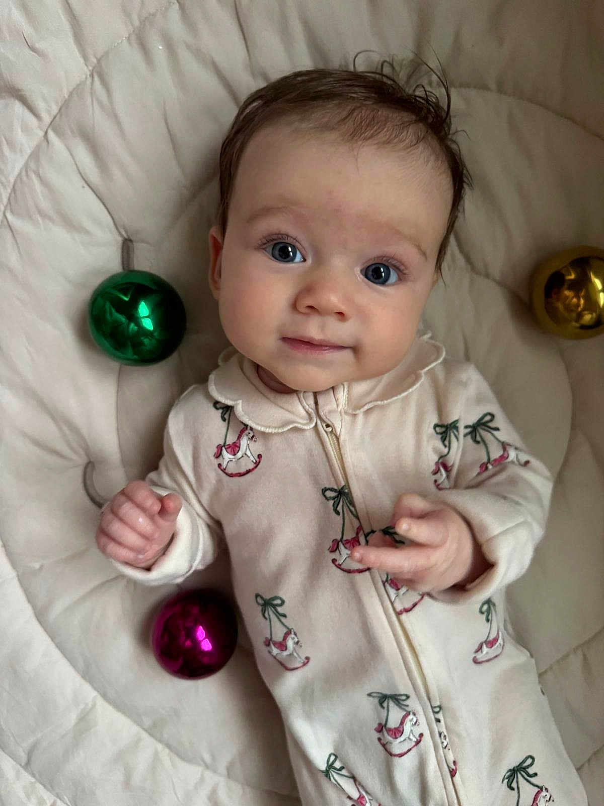 Oli is registered to the contest to win money with this photo: baby, infant, face, person, clothing, onesie, bed, quilt, ornament, christmas_bauble, green, pink, gold, expression, cute, looking_at_camera, indoor, child, small_hands, soft_lighting