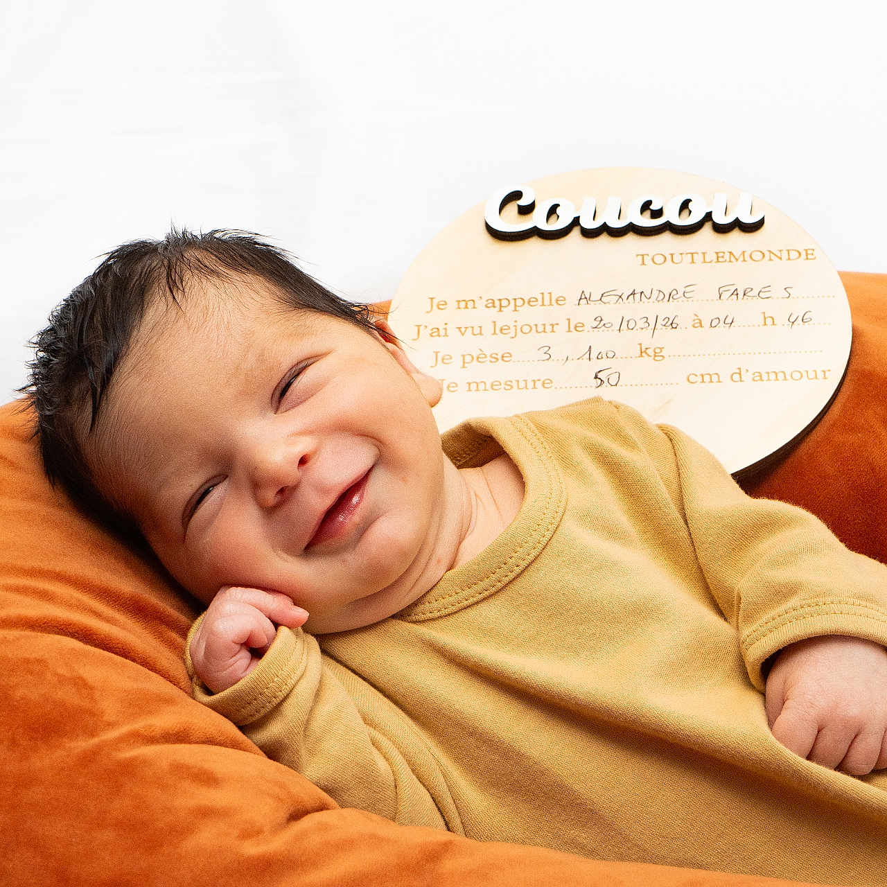 Alexandre Fares a rejoint le concours — aidez-le/la à gagner de superbes lots ! baby, newborn, smile, infant, cushion, orange, mustard, clothing, face, portrait, cute, resting, wooden_sign, text, birth_details, cozy, indoors, happy, child, person