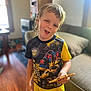 child, boy, pizza, shirt, yellow_shorts, living_room, couch, wooden_floor, smiling, holding_food, casual_clothing, indoor, furniture, person, expression, short_hair, real_photo, daylight, home, playful