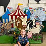 child, boy, smiling, sitting, floor, concrete, dinosaur_shirt, carnival, circus_tent, animal_cutouts, cow, horse, pig, goat, ferris_wheel, background, indoor, colorful, playful, decorations