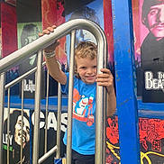 Bentlee is registered to the contest to win money with this photo: child, smiling, blue_shirt, metal_railing, outdoor, mural, colorful, portrait, happy, boy, shorts, hand, fence, music, artwork, urban, playful, person, daylight, expression