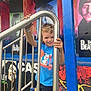 child, smiling, blue_shirt, metal_railing, outdoor, mural, colorful, portrait, happy, boy, shorts, hand, fence, music, artwork, urban, playful, person, daylight, expression