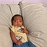 baby, blue_pants, child, clothing, couch, furniture, hand, head, indoor, infant, person, pillow, portrait, purple_wall, relaxed, serious_expression, skin, striped_pillow, wall, yellow_shirt