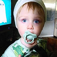 Maddox is registered to the contest to win money with this photo: toddler, child, pacifier, blue_eyes, hat, knit_hat, dinosaur_shirt, indoor, living_room, television, cartoon, ghost, toy, blurry_hand, face, portrait, person, casual_clothing, young_child, cute