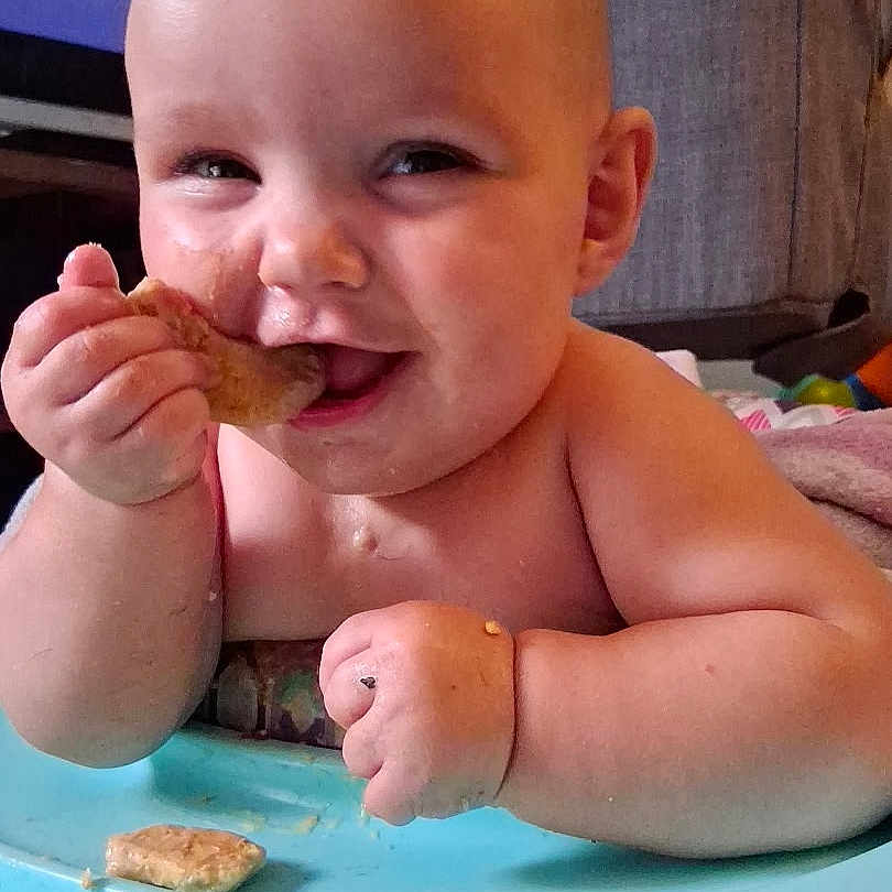 Ariel is registered to the contest to win money with this photo: baby, child, high_chair, food, eating, happy, smiling, face, hand, mouth, indoor, skin, cute, infant, mealtime, table, person, plump, cheerful, closeup