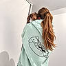 dog, dachshund, puppy, brown_dog, pet, animal, person, woman, long_hair, ponytail, hoodie, mint_green, cartoon, dog_design, indoor, portrait, shoulder, carrying, white_wall, fashion