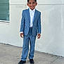 Yahir is registered to the contest to win money with this photo: boy, child, smiling, blue_suit, white_shirt, black_shoes, short_hair, standing, outdoor, concrete, sidewalk, wall, window, formal_wear, portrait, person, young, full_body, casual_background, happy