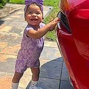 Kataleia is registered to the contest to win money with this photo: toddler, child, smiling, purple_bow, romper, sneakers, socks, car, red_car, outdoor, sunny, green_grass, patio, tiles, happy, standing, cute, playful, young_child, daylight