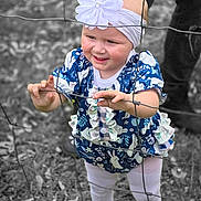 Ellie is registered to the contest to win money with this photo: toddler, child, girl, headband, flower, dress, wire_fence, curious, outdoor, grass, leaves, legs, adult, white_shoes, playful, color_pop, black_and_white_background, nature, standing, portrait