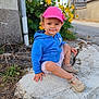 blue_hoodie, cap, casual_clothing, child, concrete_curb, daylight, greenery, happy, nature, outdoor, pink_cap, plants, rural, shoes, sidewalk, sitting, smiling, street, toddler, yellow_flowers