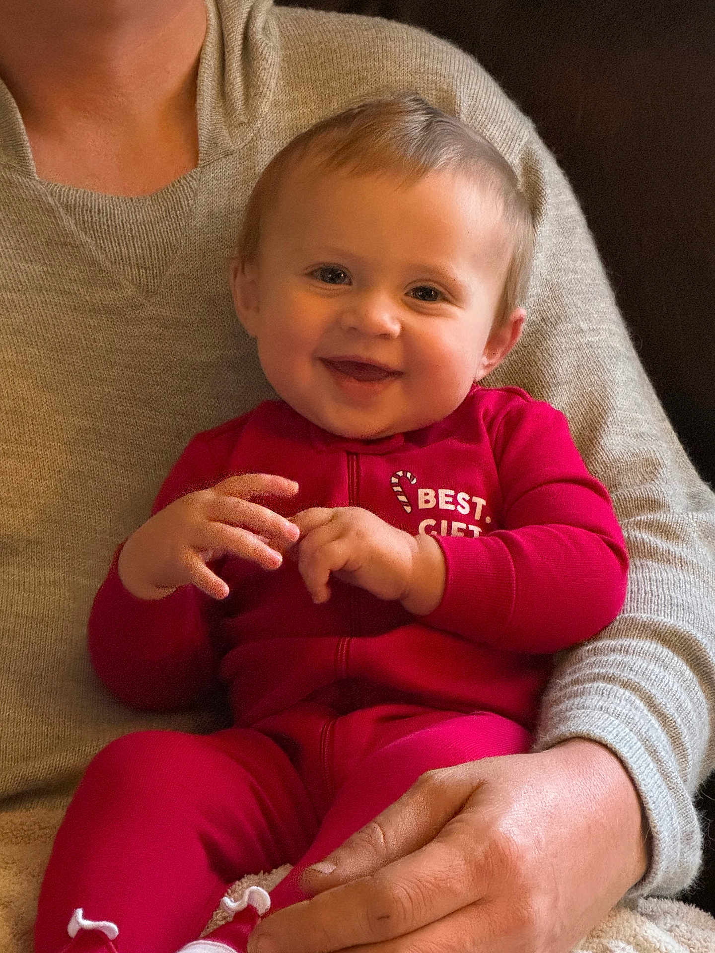 Riley is registered to the contest to win money with this photo: baby, child, smiling, red_onesie, person, adult_hand, cozy, indoor, cute, happy, festive, holiday, candid, portrait, warm_lighting, infant, sitting, clothing, face, human