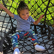 Daniel is registered to the contest to win money with this photo: child, smiling, braided_hair, net_swing, outdoor, grass, blue_shirt, colorful_leggings, white_socks, happy, playful, daylight, young_child, woven_swing, sunny, park, recreation, seated, person, nature