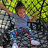 child, smiling, braided_hair, net_swing, outdoor, grass, blue_shirt, colorful_leggings, white_socks, happy, playful, daylight, young_child, woven_swing, sunny, park, recreation, seated, person, nature