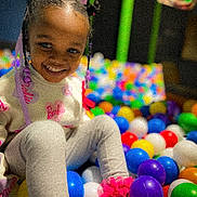 Gianna is registered to the contest to win money with this photo: child, smiling, ball_pit, colorful, braided_hair, pink_beads, playground, happy, indoor, toy, legs, sitting, casual_clothing, young, cute, fun, leisure, person, play, entertainment