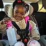 child, girl, car_seat, water_bottle, braids, red_beads, coat, heart_pattern, pants, smile, seatbelt, car_interior, safety, cute, portrait, warm_clothing, happy, young_child, holding_object, indoors