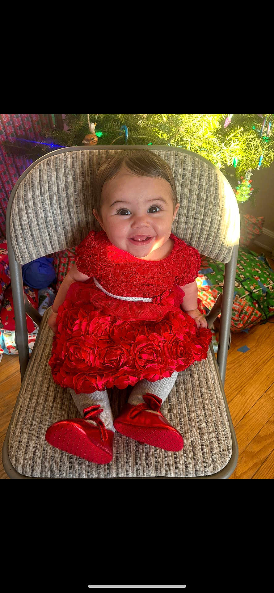 Alaia is registered to the contest to win money with this photo: baby, child, smiling, red_dress, chair, christmas_tree, presents, holiday, indoors, happy, cute, festive, floor, decorations, toy, person, portrait, toddler, footwear, warm_lighting