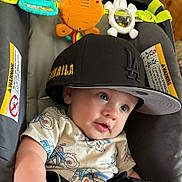 Jameson is registered to the contest to win money with this photo: baby, child, hat, cap, black, tshirt, toy, seat, infant, blue_eyes, face, cute, indoor, clothing, pattern, expression, person, comfort, play, closeup
