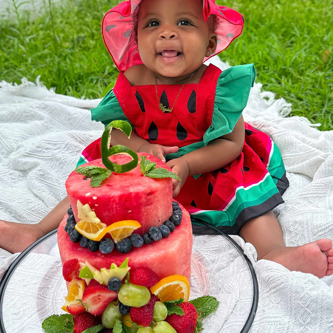 Azalea is registered to the contest to win money with this photo: baby, blanket, blueberries, celebration, child, cute, fence, fruit_cake, grapes, greenery, happy, hat, orange_slices, outdoor, portrait, sitting, smiling, strawberries, watermelon_cake, watermelon_dress