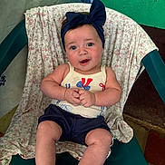Luna is registered to the contest to win money with this photo: baby, child, smiling, headband, bow, sitting, chair, clothing, tank_top, shorts, indoor, fabric, pattern, green_wall, floor, person, cute, happy, portrait, skin