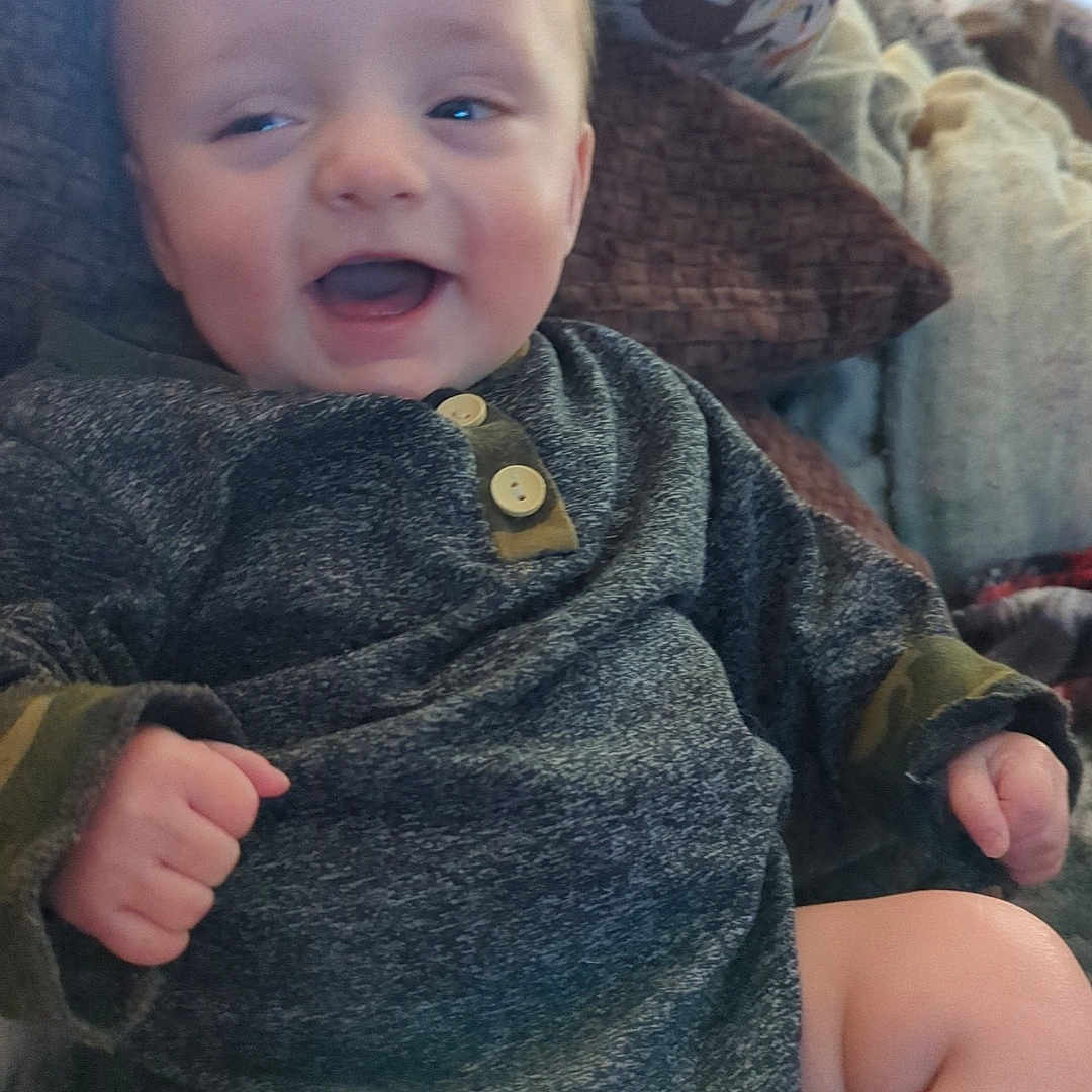 Colt is registered to the contest to win money with this photo: baby, bodypart, clothing, coat, couch, cushion, face, finger, furniture, hand, happy, head, jacket, knitwear, pants, person, photography, portrait, sitting, sweater