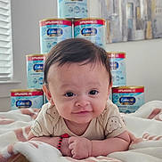 Matias is registered to the contest to win money with this photo: baby, infant, blanket, formula_cans, indoor, smiling, face, arm, person, child, furniture, window, blinds, chair, portrait, cute, lying_down, expression, room, soft_lighting