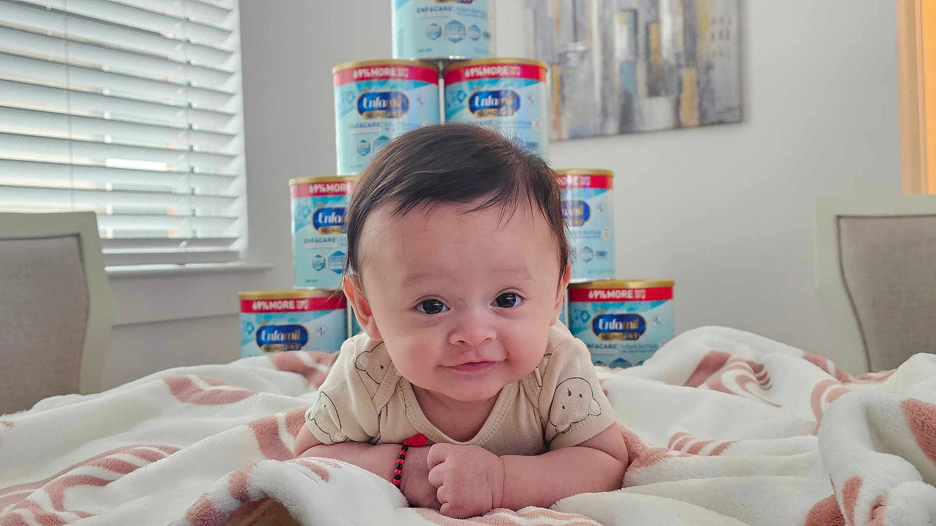 Matias is registered to the contest to win money with this photo: baby, infant, blanket, formula_cans, indoor, smiling, face, arm, person, child, furniture, window, blinds, chair, portrait, cute, lying_down, expression, room, soft_lighting