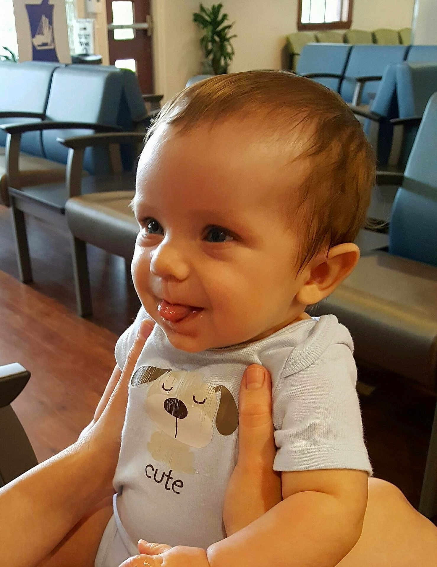 Isaac Lasserre is registered to the contest to win money with this photo: baby, child, tongue_out, onesie, cute, smiling, hands, indoor, waiting_room, chairs, wood_floor, person, infant, short_sleeves, holding, face, expression, young, light, casual