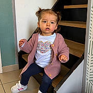 Jade participe au concours pour gagner de l'argent avec cette photo : child, toddler, pigtails, pink_cardigan, heart_tshirt, sneakers, sitting, staircase, wooden_stairs, tiled_floor, indoor, curious_expression, portrait, young_child, buttons, blue_leggings, white_shoes, wall, doorframe, cozy
