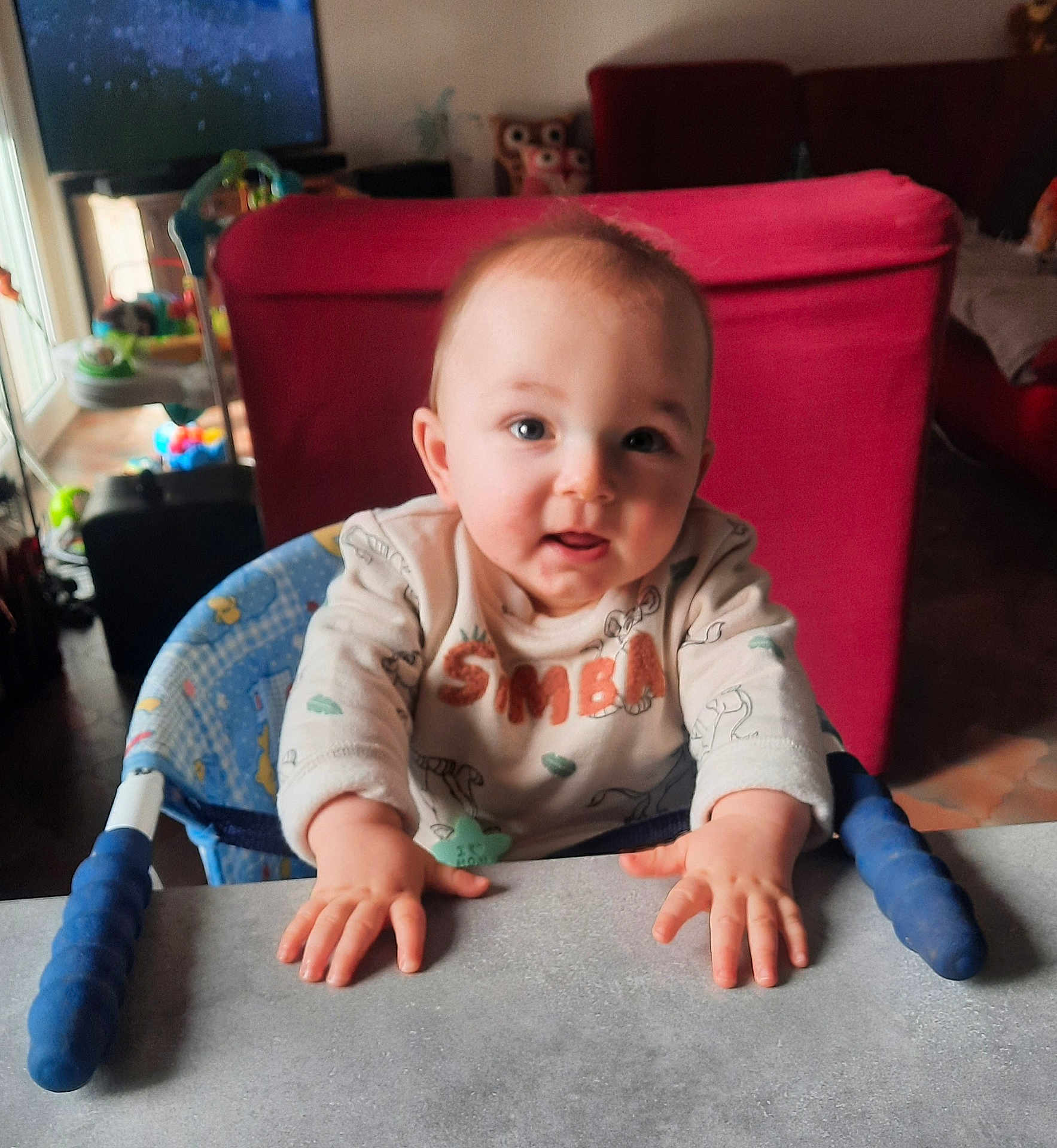 Theo participe au concours pour gagner de l'argent avec cette photo : baby, child, infant, smiling, hands, table, highchair, indoor, living_room, red_couch, toy, face, person, cute, young, clothing, expression, curious, home, furniture
