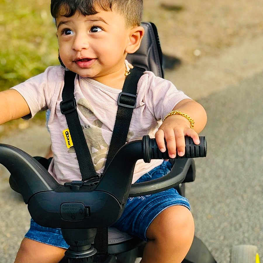 Adwick joined the competition — help win amazing prizes! black_tricycle, bracelet, child, curly_hair, expression, foot, grass, handlebars, happy, outdoor, pavement, playtime, seatbelt, shirt, shorts, smiling, summer_clothing, toddler, tricycle, young_child
