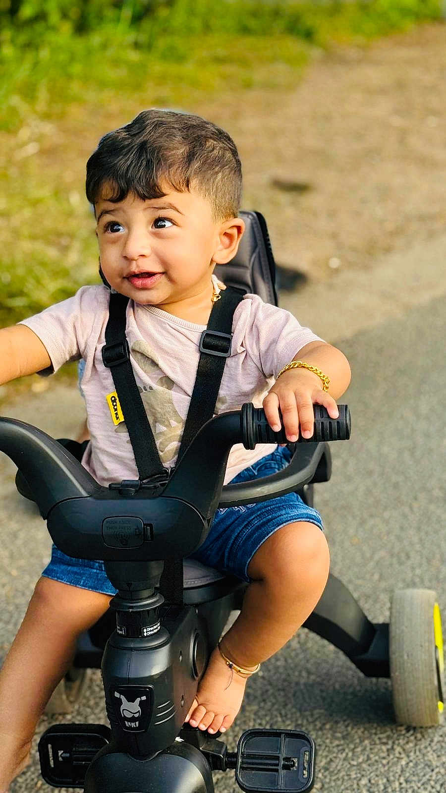 Adwick joined the competition — help win amazing prizes! toddler, child, tricycle, outdoor, smiling, shorts, shirt, grass, pavement, handlebars, foot, bracelet, black_tricycle, happy, young_child, summer_clothing, playtime, seatbelt, expression, curly_hair