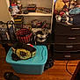 cat, basket, stuffed_animals, shelf, dvds, toy, plastic_bin, championship_belt, slippers, wood_floor, drawer, blanket, remote_control, indoor, furniture, small_cat, plush_toy, wire_basket, clutter, home