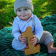 Frankie is registered to the contest to win money with this photo: baby, child, outdoor, grass, blanket, hat, knitted_hat, pom_pom, number_one, toy, smile, happy, cute, portrait, sitting, clothing, green, sunlight, face, hands