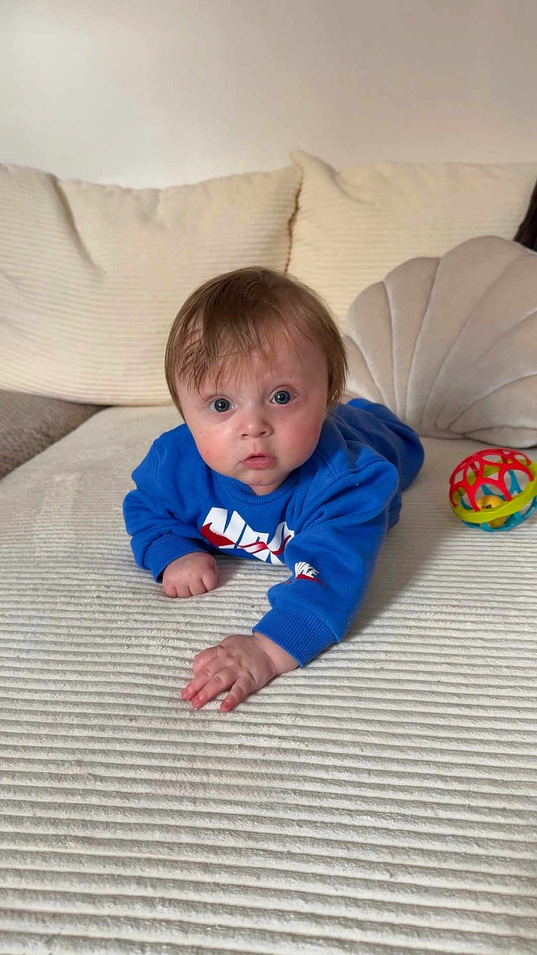 Leandre a rejoint le concours — aidez-le/la à gagner de superbes lots ! baby, blue_sweatshirt, curious, indoors, couch, toy_ball, child, face, hand, expression, fabric, texture, home, young_child, person, sitting, playful, comfortable, soft_lighting, closeup