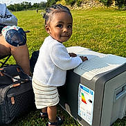 Meera a rejoint le concours — aidez-le/la à gagner de superbes lots ! toddler, child, outdoor, grass, cooler, white_sweater, striped_skirt, sandals, nike, dimples, braided_hair, picnic, sunny, trees, bag, person, legs, nature, daytime, park