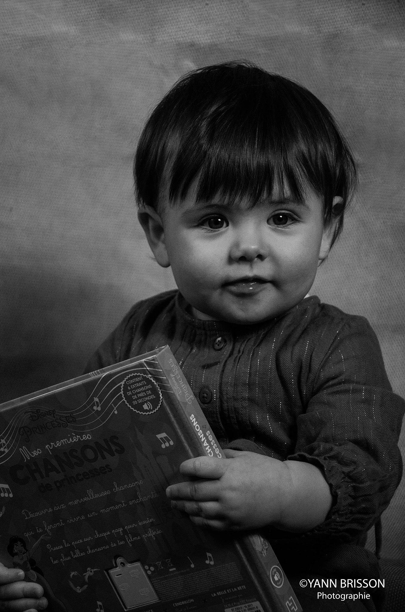 Judith participe au concours pour gagner de l'argent avec cette photo : bangs, black, child, darkness, flash_photography, happy, human_body, monochrome, monochrome_photography, person, photography, portrait, portrait_photography, reading, room, sitting, smile, standing, toddler, toy