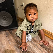 Azarion is registered to the contest to win money with this photo: baby, child, floor, wooden_floor, speaker, wall, curious, sitting, hands, clothing, brown_skin, indoor, expression, young, portrait, cute, crawling, sock, black_hair, room