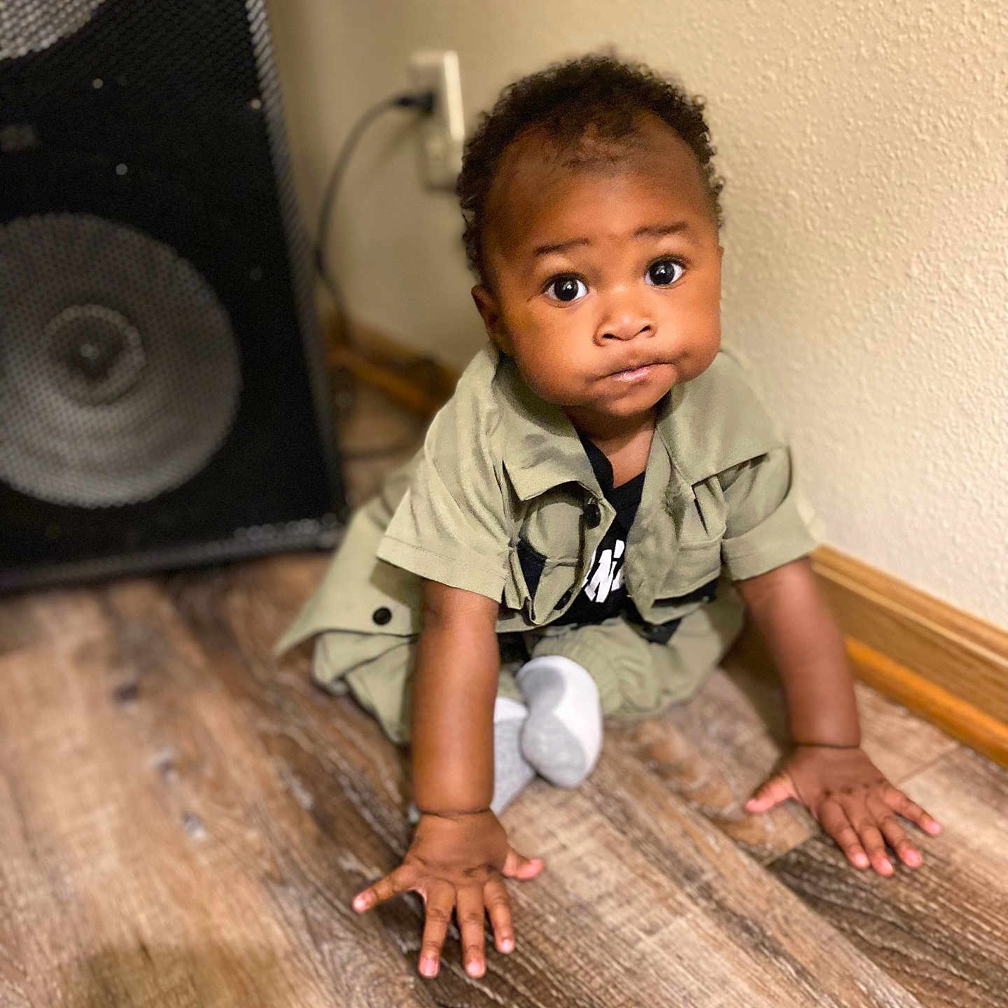 Azarion is registered to the contest to win money with this photo: baby, black_hair, brown_skin, child, clothing, crawling, curious, cute, expression, floor, hands, indoor, portrait, room, sitting, sock, speaker, wall, wooden_floor, young