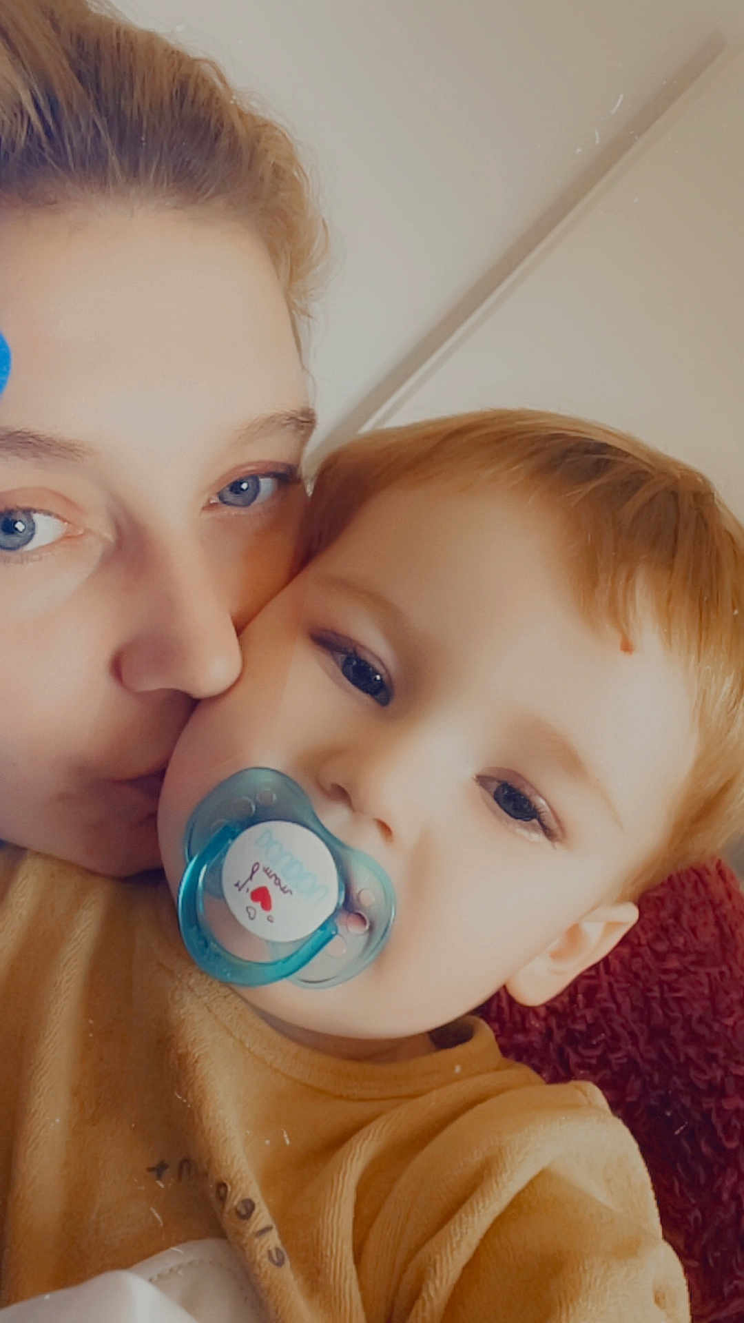 Maël participe au concours pour gagner de l'argent avec cette photo : adult, child, toddler, pacifier, kiss, face, closeup, indoor, love, affection, blue_eyes, blond_hair, yellow_clothing, blanket, cuddle, cozy, person, portrait, rest, comfort